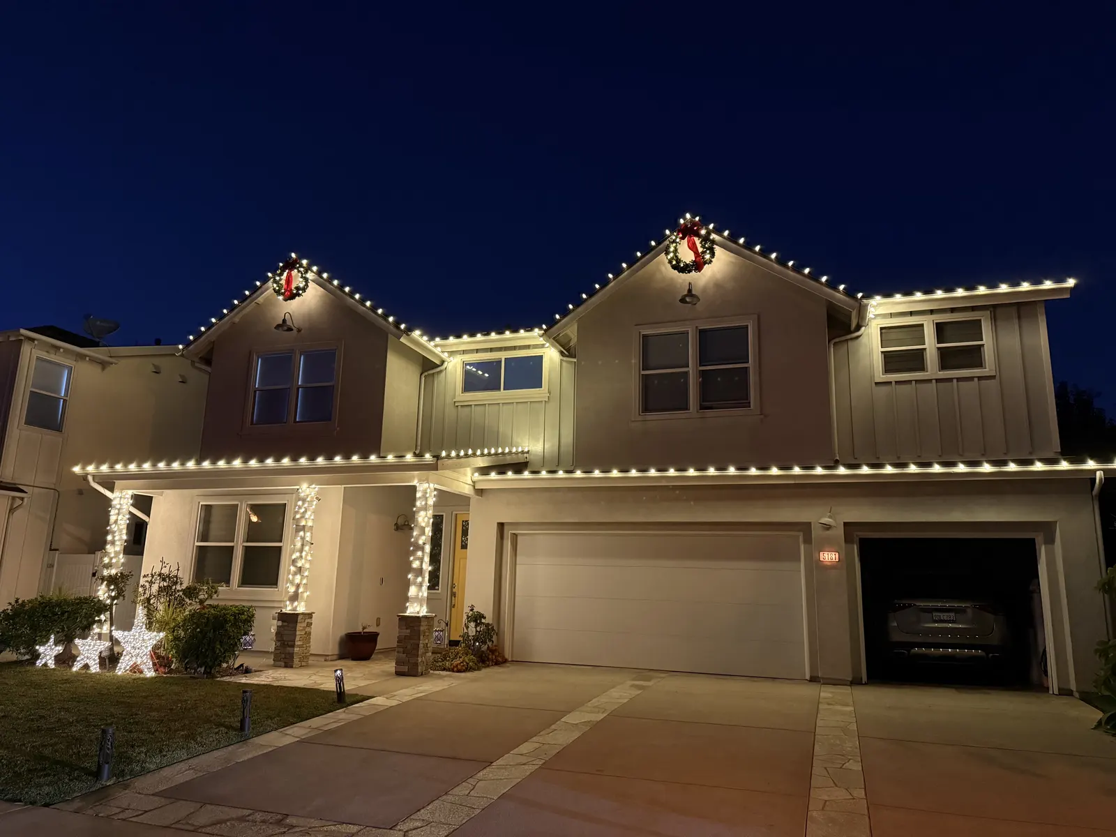 Two-story home with warm white Christmas lights outlining every roof peak and a wreath above the entry