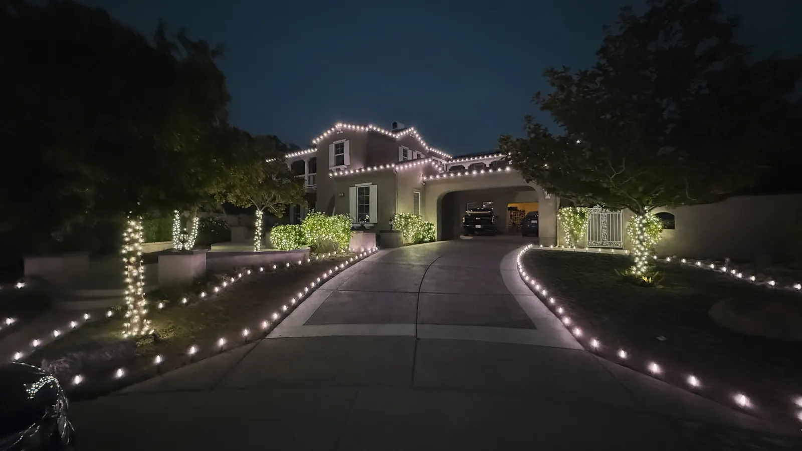 Long driveway lined with Christmas lights leading to a lit home with wrapped trees