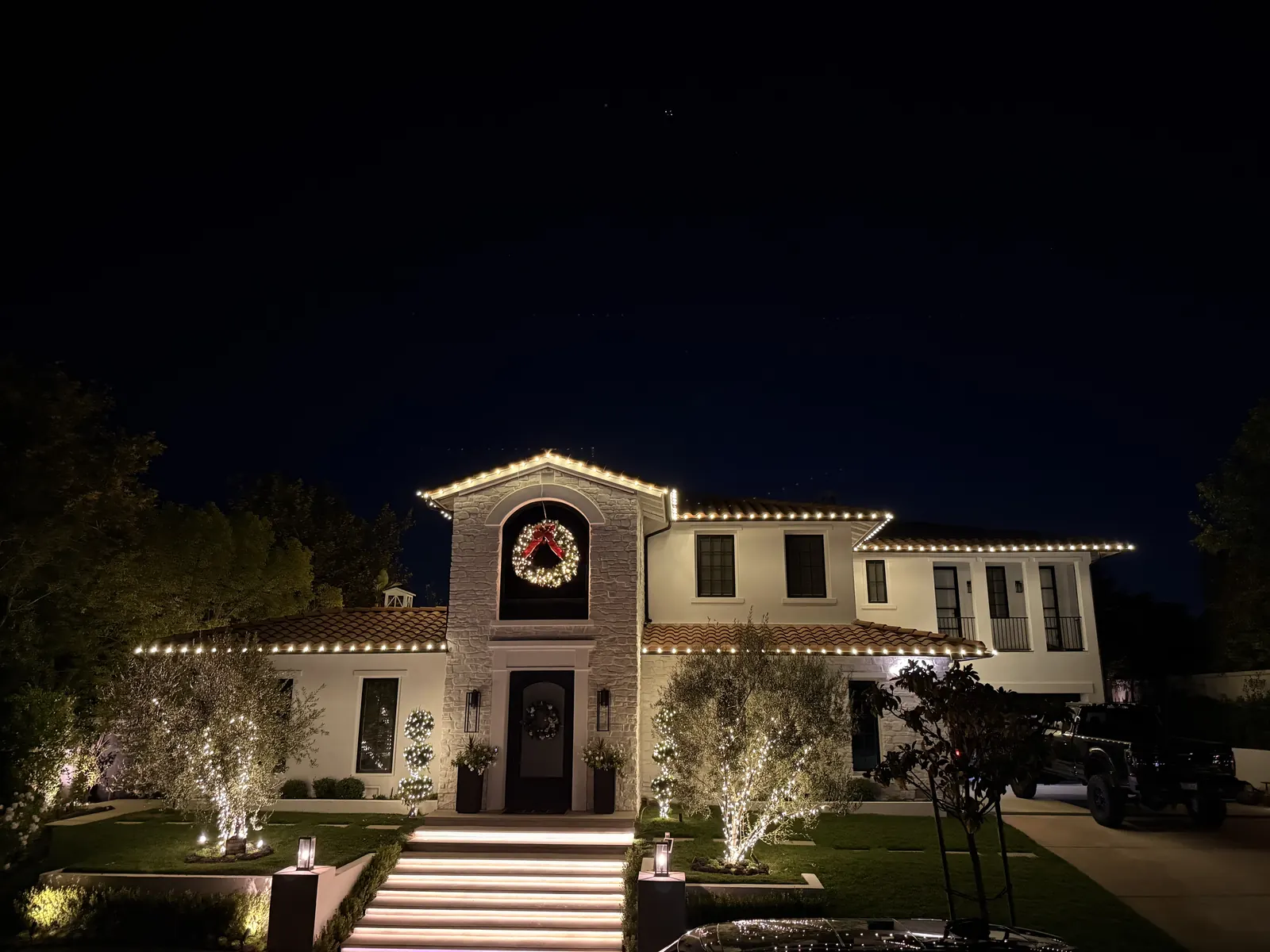 Two-story home in La Cañada Flintridge with warm white roofline lights, a lit wreath, and uplit trees by the front steps