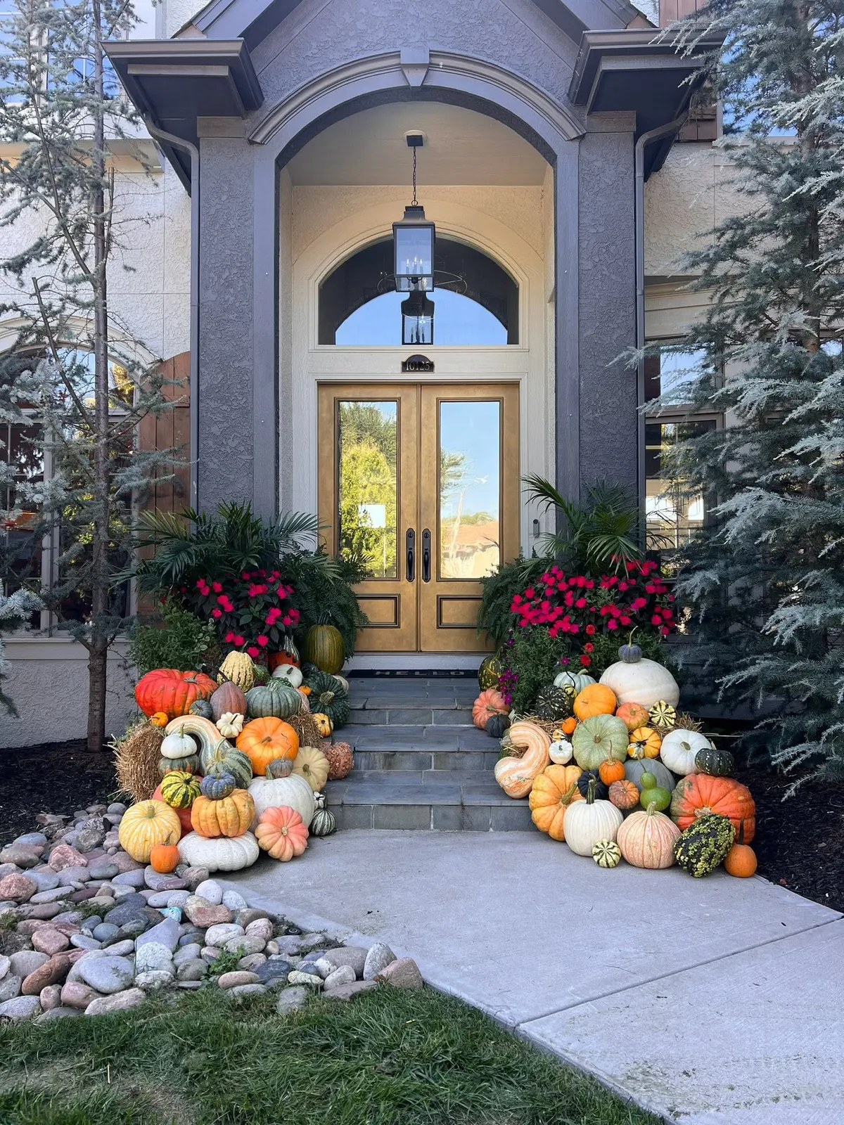 Large mix of heirloom, white, and orange pumpkins flanking a gray arched home entrance with red flowers