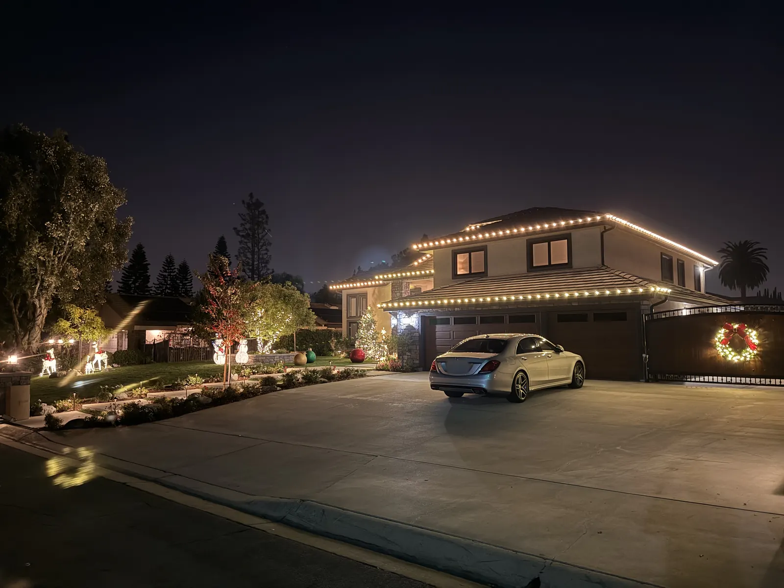 Two-story home in Yorba Linda with warm white roofline lights, a lit wreath on the driveway gate, and holiday decorations on the lawn